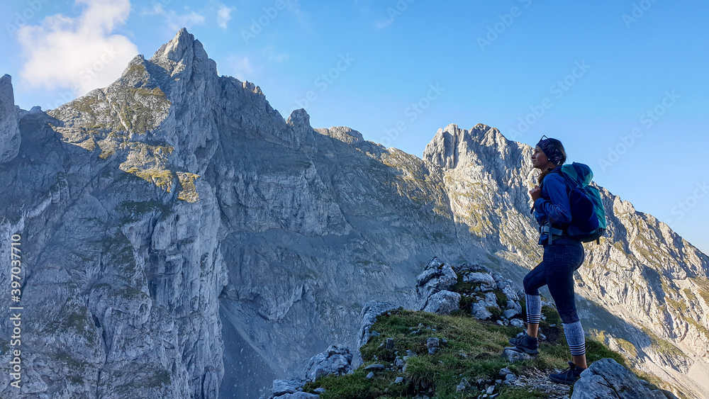 A woman with a backpack standing in the shadow at the mountain peak and enjoying the view on higher, very steep peaks in front of him. Way to the Grimming in Austrian Alps. Freedom and exploration