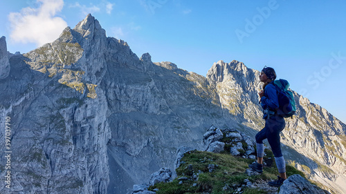 A woman with a backpack standing in the shadow at the mountain peak and enjoying the view on higher, very steep peaks in front of him. Way to the Grimming in Austrian Alps. Freedom and exploration