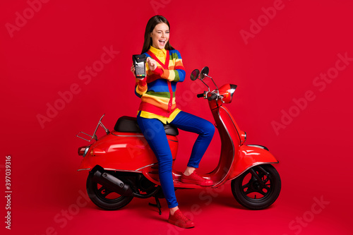 Photo portrait of winking girl paying via terminal with credit card sitting on scooter isolated on vivid red colored background
