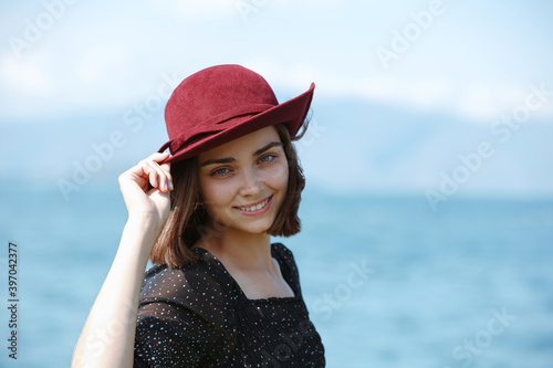 Woman smiling and touching hat while standing against blue sea and looking at camera enjoying summer holiday  © Dzmitry