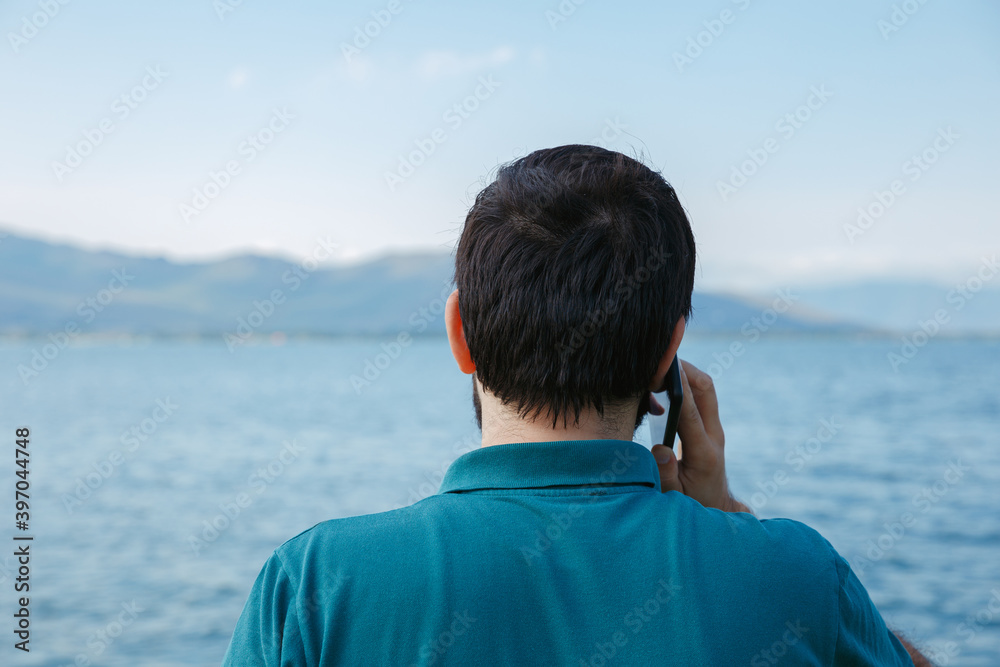 From back view man talking on smartphone and standing on background of blue sea in summer during vacation From back view man talking on smartphone and standing on background of blue sea in summer during vacation