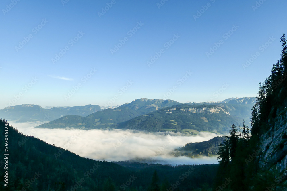 A panoramic view on an Alpine valley covered with a thick layer of fog. High mountain peaks popping above the fog. A few trees on the sides.  Early morning haze in the Austrian Alps. Expedition