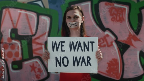 WE WANT NO WAR - young caucasian brunette female activist with typed mouth on the street against the graffiti wall protests against the war. Political issues. Young people demand peaceful future, 4k photo