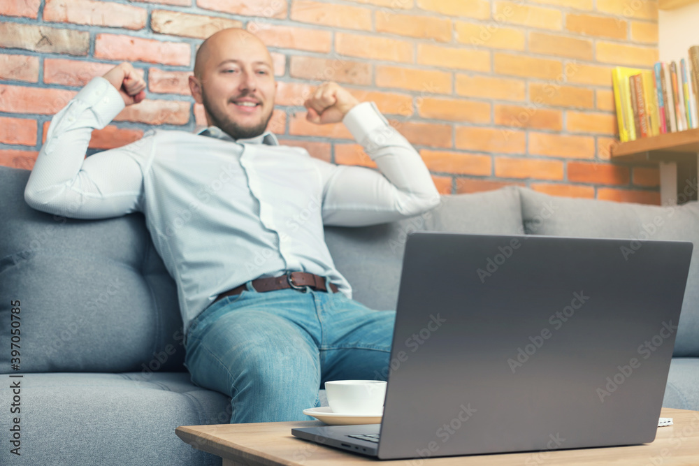 Defocused bearded bald man, businessman or freelancer sitting on sofa, has happiness emotions and winner feeling, working on laptop from home, modern interior loft design
