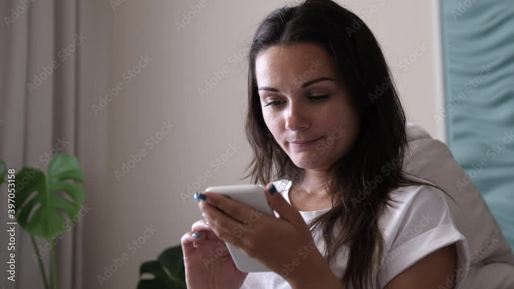 Happy young woman holding smart phone looking at cellphone screen enjoying using mobile apps for shopping having fun playing games chatting in social media sit on couch at home Happy young woman holding smart phone looking at cellphone screen enjoying using mobile apps for shopping having fun playing games chatting in social media sit on couch at home