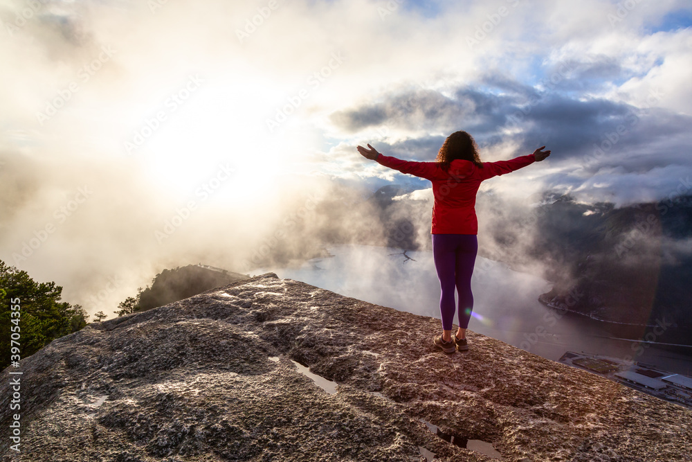 Adventurous Girl Hiking on top of a Peak, Chief Mountain, during a dramatic colorful sunny sunset. Located in Squamish, North of Vancouver, British Columbia, Canada.