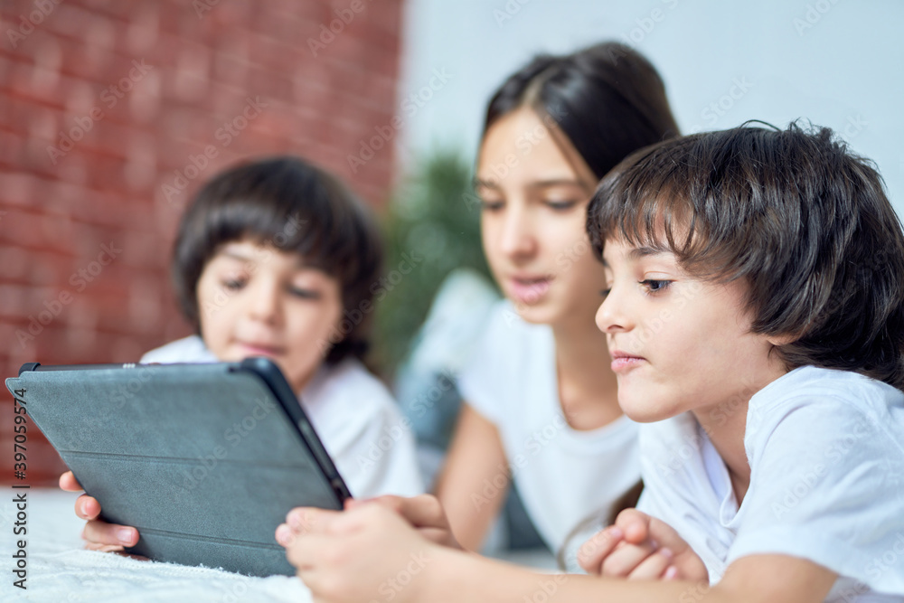 Curious little latin boy looking at the screen of tablet pc. Brother spending time with his siblings, playing games, lying on the bed at home Curious little latin boy looking at the screen of tablet pc. Brother spending time with his siblings, playing games, lying on the bed at home