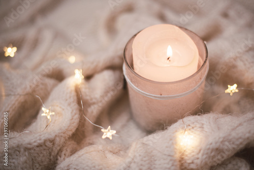 A candle shines in a candlestick that stands on a beige knitted blanket. Christmas garland near.