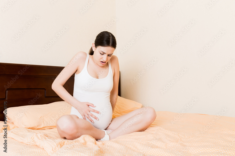 Young pretty pregnant woman, in pain, sitting on bed in bedroom, preparing to give a birth Young pretty pregnant woman, in pain, sitting on bed in bedroom, preparing to give a birth