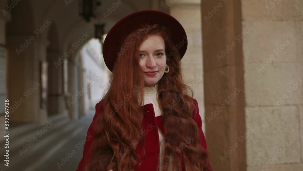 Fashionable young happy smiling redhead woman wearing red coat and hat touching her curly hair, elegant ginger girl. Posing at street of European city. Autumn, winter fashion. Slow Motion 100 fps Fashionable young happy smiling redhead woman wearing red coat and hat touching her curly hair, elegant ginger girl. Posing at street of European city. Autumn, winter fashion. Slow Motion 100 fps