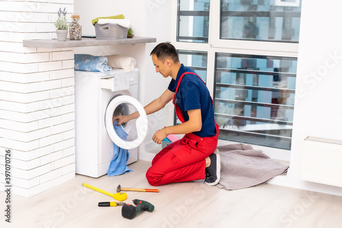 Plumber in overalls with tools is repairing a washing machine in the house