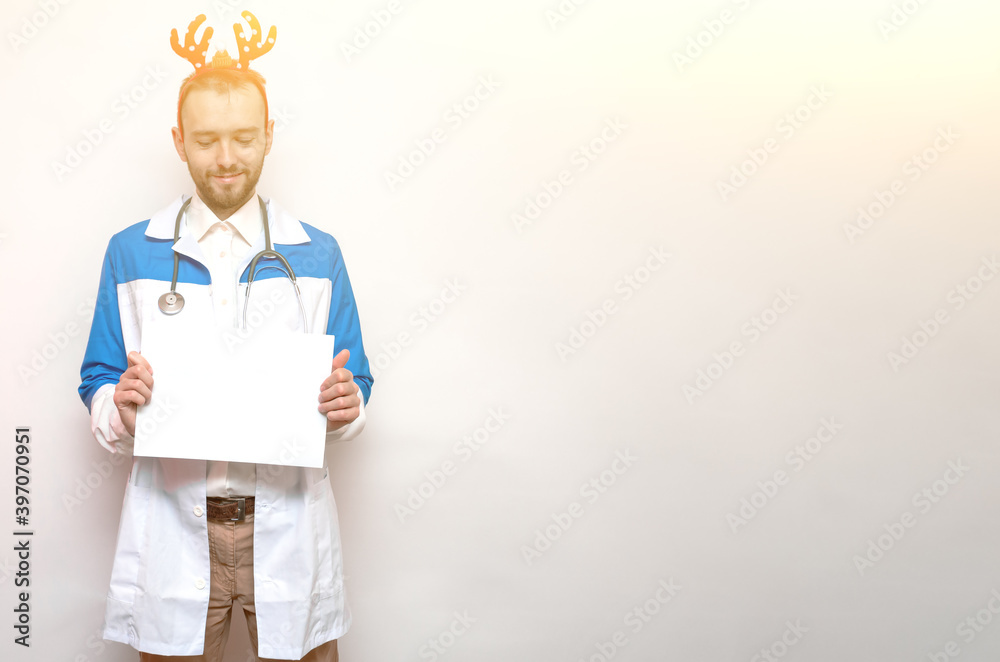 Young bearded doctor with toy deer antlers holds a blank piece of paper in his hands on a white background. Isolated. Christmas and New Year concept. Portrait. Copy space. Place for text Young bearded doctor with toy deer antlers holds a blank piece of paper in his hands on a white background. Isolated. Christmas and New Year concept. Portrait. Copy space. Place for text