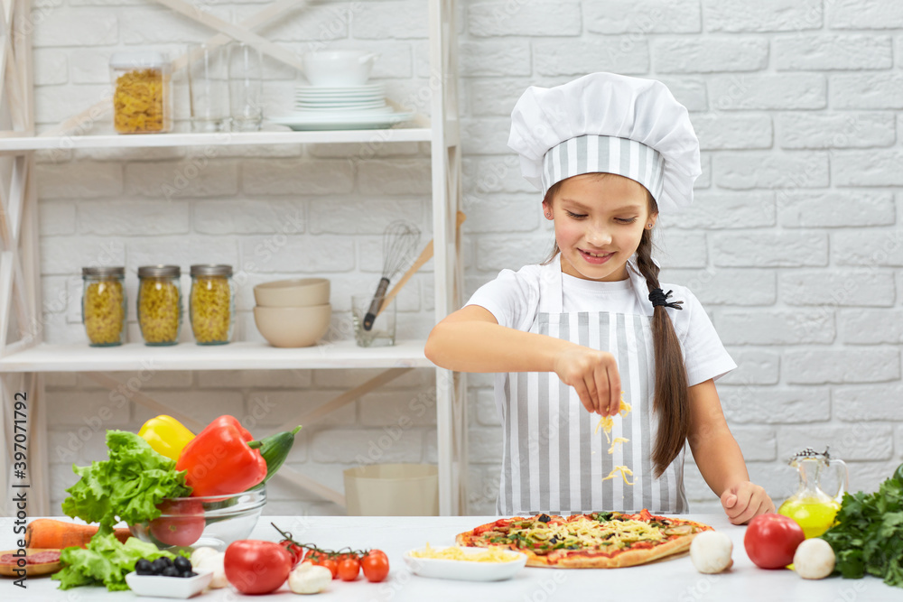 little girl in chef hat and an apron cooking pizza in the kitchen. child adding cheese into pizza little girl in chef hat and an apron cooking pizza in the kitchen. child adding cheese into pizza