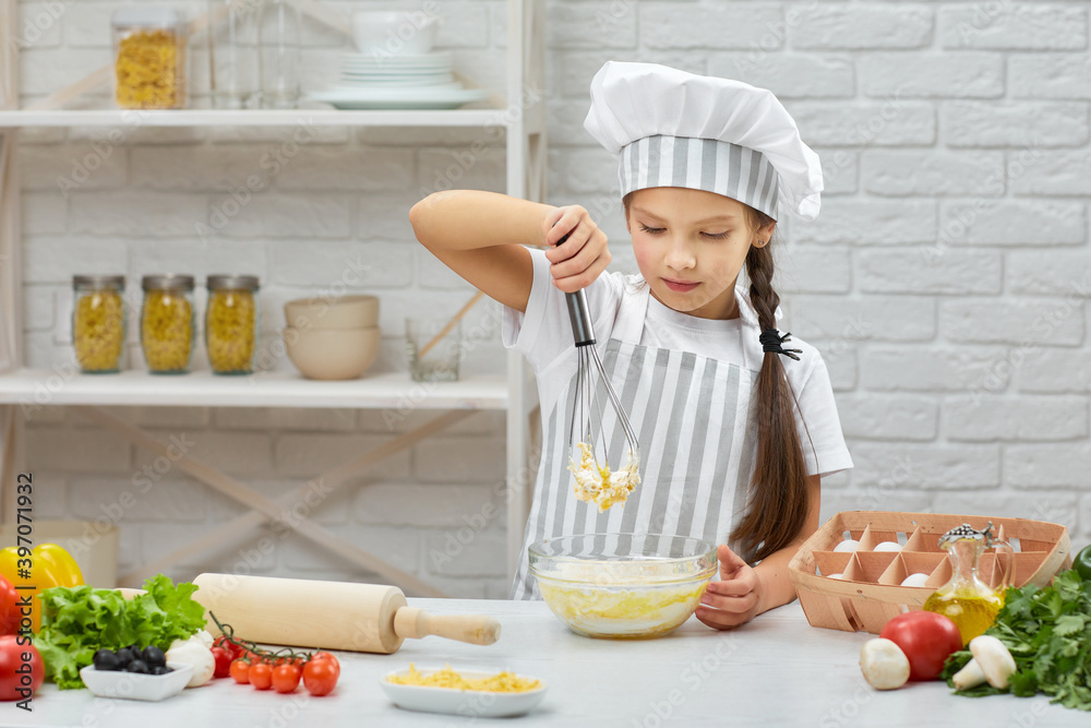 little child girl in cap and an apron kneads with corolla the dough in the kitchen. Whisking. little child girl in cap and an apron kneads with corolla the dough in the kitchen. Whisking.