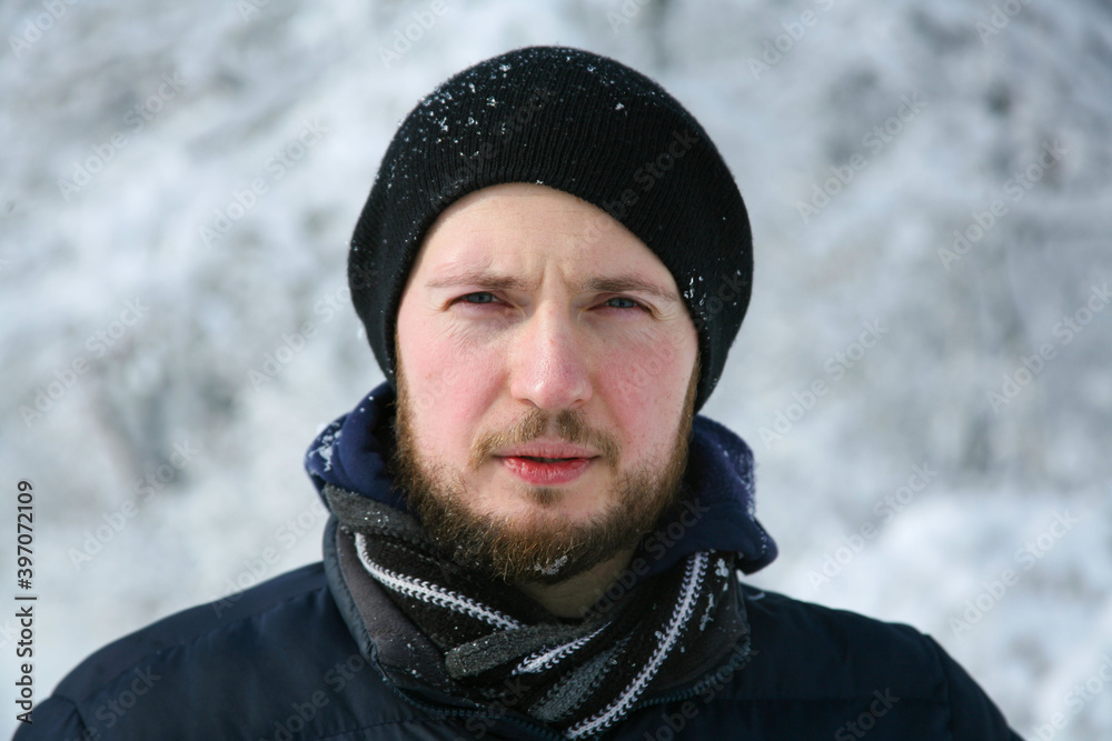 A young man in warm winter clothes is looking at the camera. A man in the winter in the forest. Portrait of a man with a beard