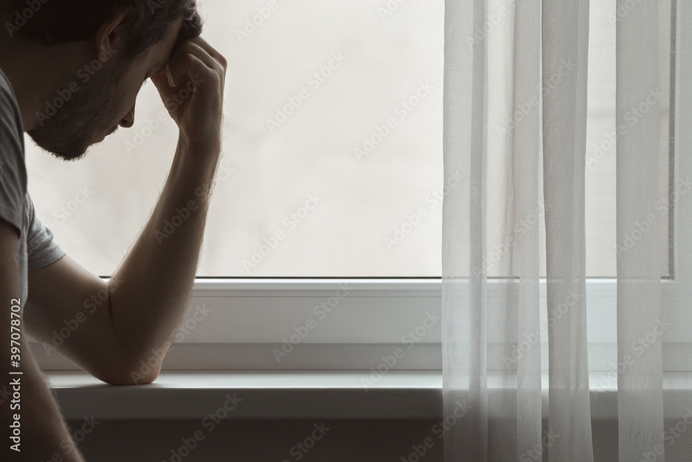 One sad serious young adult man sitting at window in home. Touching head with hand. Thinking about life. Side view. Dark gloomy atmosphere. One sad serious young adult man sitting at window in home. Touching head with hand. Thinking about life. Side view. Dark gloomy atmosphere.