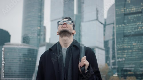 Man in coat looking up to the sky on background of skyscrapers pan shot right to left. Gimbal shot of businessman in eyeglasses near skyscrapers in business city