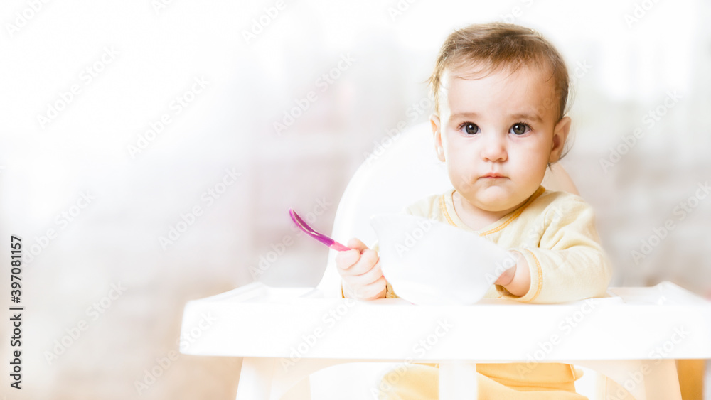 Little baby sitting in high chair and eating food. baby's first feeding. 6 month old toddler holding spoon and plate Little baby sitting in high chair and eating food. baby's first feeding. 6 month old toddler holding spoon and plate
