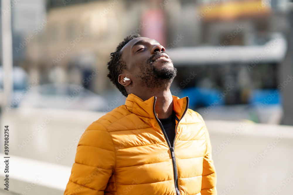 African man listening to music and breathing fresh air outdoors standing in the city - obrazy, fototapety, plakaty 