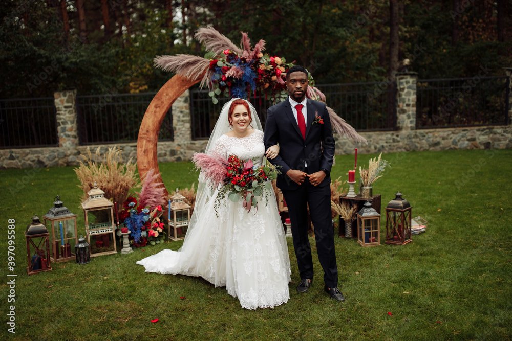 Charming African American groom with gorgeous white wife stand near decorated arch, lovely woman with handsome man on wedding ceremony, smiling, happy marriage concept