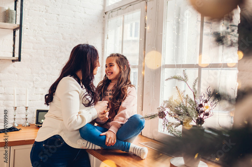 Mother and daughter relaxing on kitchen window sill by decorated Christmas tree. Family having fun together at home