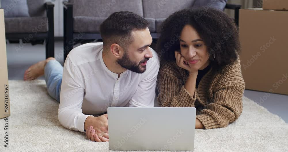 Close-up multiethnic couple curly black woman and caucasian man lie on carpet on floor of house living room use laptop to communicate talking to webcam online speaking to friends greeting waves hand Close-up multiethnic couple curly black woman and caucasian man lie on carpet on floor of house living room use laptop to communicate talking to webcam online speaking to friends greeting waves hand