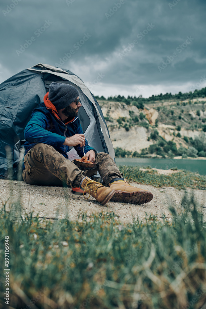 Young attractive bearded man sitting next to a tent on a camping trip and preparing lunch. Young attractive bearded man sitting next to a tent on a camping trip and preparing lunch.