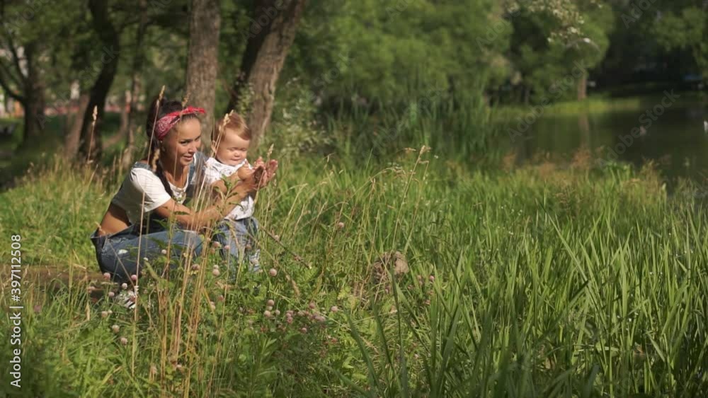 Mother with baby son sitting near pond in park. Toddler trying to clap hands together with mom, looking around. Child experiencing beauty of nature: green trees, birds and water. Concept of motherhood Mother with baby son sitting near pond in park. Toddler trying to clap hands together with mom, looking around. Child experiencing beauty of nature: green trees, birds and water. Concept of motherhood