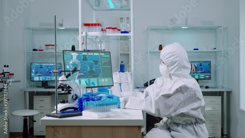 Woman biochemist in coverall checking manifestations of virus working on comuper in equipped lab. Team of doctors examining vaccine evolution using high tech researching diagnosis against covid19 photo