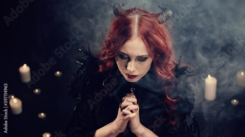 Red-haired girl witch with black horns sitting on floor in candlelight and praying with folded hands. Scary demon doing witchcraft. Halloween mystery rite. Ritual of Occultism or esoterics concept.