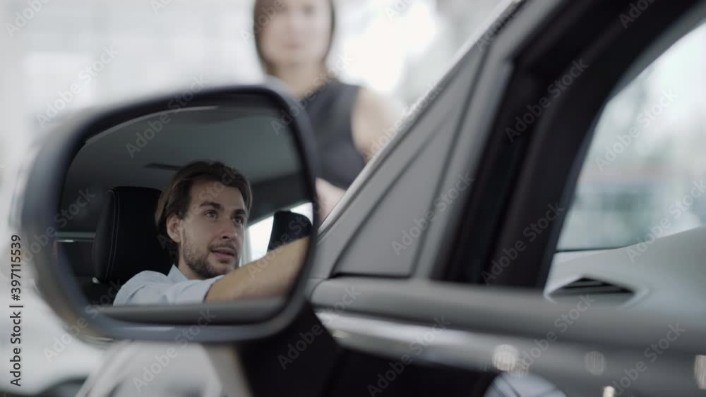 Reflection in car side mirror of smiling handsome man listening to blurred woman talking at the background. Happy Caucasian client sitting in new vehicle in car dealership. Automobile industry. Reflection in car side mirror of smiling handsome man listening to blurred woman talking at the background. Happy Caucasian client sitting in new vehicle in car dealership. Automobile industry.