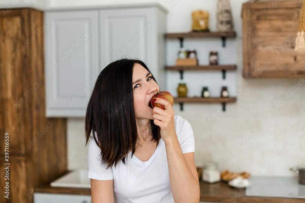 Brunette girl in a white T-shirt in the kitchen eat an apple. Brunette girl in a white T-shirt in the kitchen eat an apple.