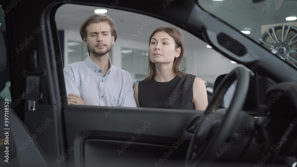 Young confident couple examining new vehicle in car dealership or showroom and talking. Portrait of successful Caucasian man and woman choosing automobile. Shooting from salon. Young confident couple examining new vehicle in car dealership or showroom and talking. Portrait of successful Caucasian man and woman choosing automobile. Shooting from salon.