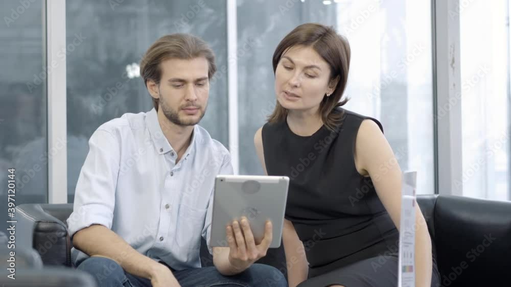 Concentrated Caucasian man and woman using tablet and talking. Portrait of elegant successful colleagues or partners discussing business deal indoors. Lifestyle and business concept. Concentrated Caucasian man and woman using tablet and talking. Portrait of elegant successful colleagues or partners discussing business deal indoors. Lifestyle and business concept.