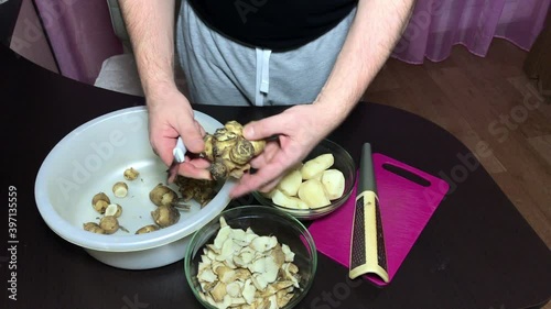 A man peels Jerusalem artichoke with a kitchen knife. Throws the peel into one container, the peeled vegetable into another.