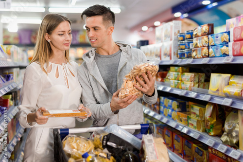 Young glad positive loving couple making purchases together, choosing cookies in grocery store