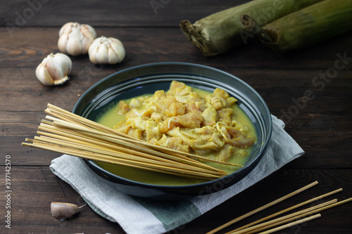 Preparation for making Sate Ayam or Indonesian Chicken Satay, chicken slices marinated on grey ceramic bowl, with rice cake and bamboo skewer. Dark wooden background.  photo