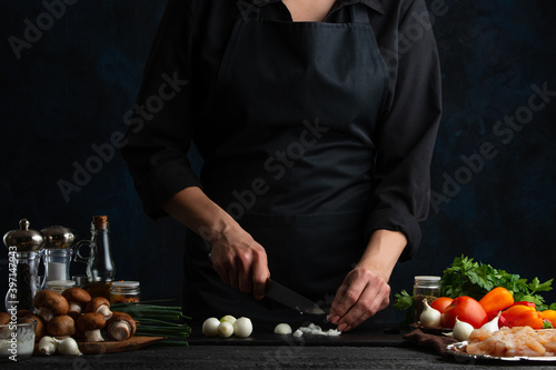 Professional chef in black uniform cuts with knife white onion. Concept of cooking process. Backstage of preparing meat filling for pie on dark blue background. Frozen motion.
