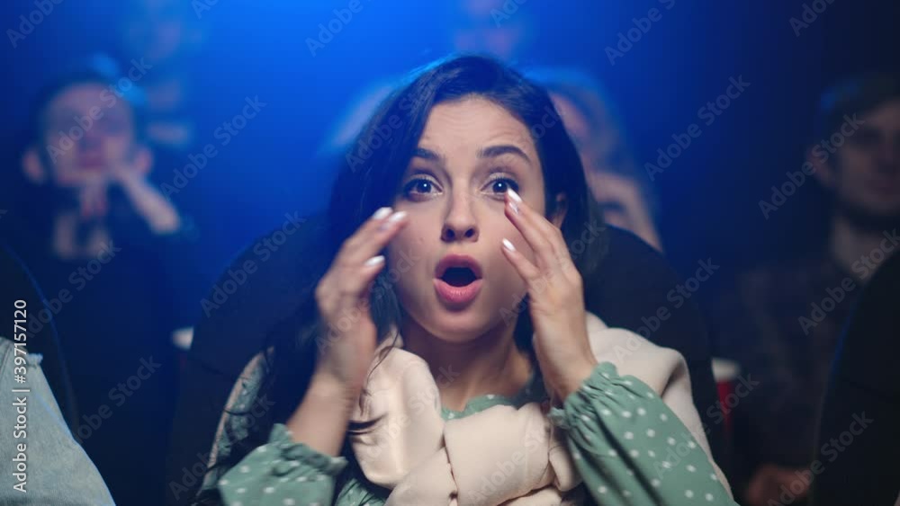 Shocked woman watching scary movie in dark hall. Scared girl eating in cinema.