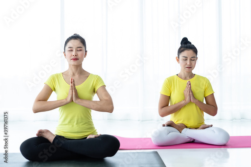Two young Asian women workout practicing yoga in yellow dress or pose with a trainer and practice meditation wellness lifestyle and health fitness concept in a gym. © Thinapob