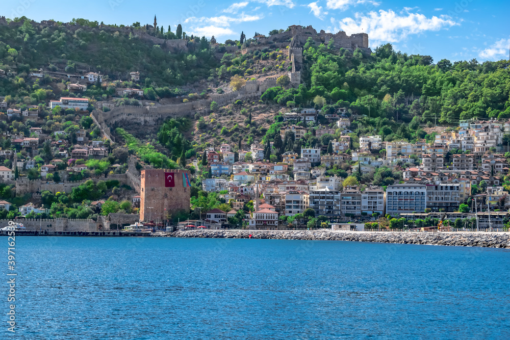 View from the sea to the Old City of Alanya (Turkey) with the Red Tower on the background of turquoise water. Panorama of a town with modern buildings on the hillside and an ancient castle at the top