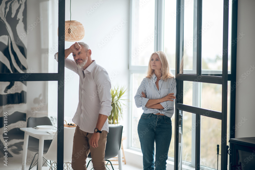 Bearded man looking sad, blonde woman watching him with arms crossed