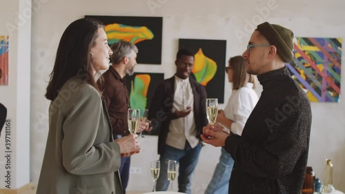 Young man and woman holding flutes with champagne and discussing something at exhibition opening party in art gallery photo