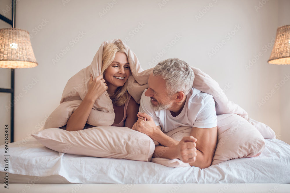 Grey-haired man and a blonde woman lying in bed and having fun Grey-haired man and a blonde woman lying in bed and having fun