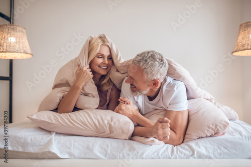 Grey-haired man and a blonde woman lying in bed and having fun