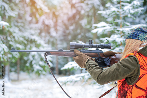 A young beautiful hunter woman in camouflage with rifle ready for hunt in winter landscape.