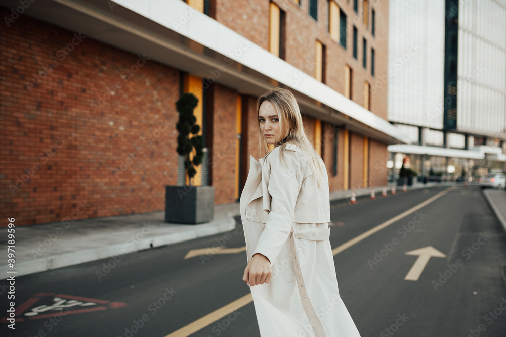 Serious trendy young girl in a beige trench coat in an elegant jeans in trench coat posing outdoors on a road. 