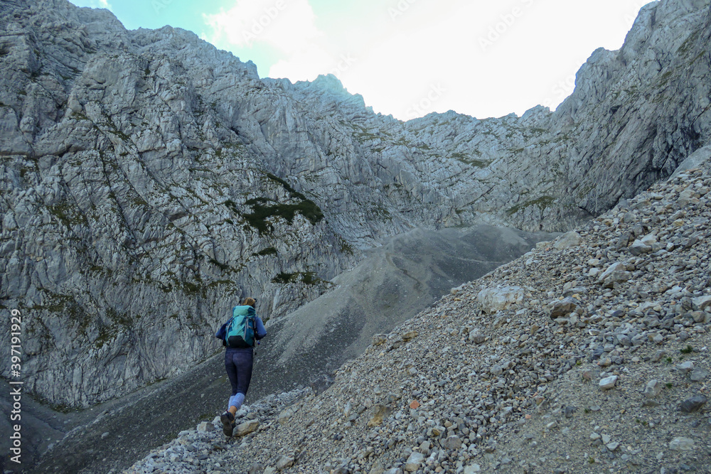 A woman with backpack hiking through steep and high mountain crater in Austrian Alp. The sun shines behind the mountain wall. The whole area is covered with shadow. Steep and narrow path along.
