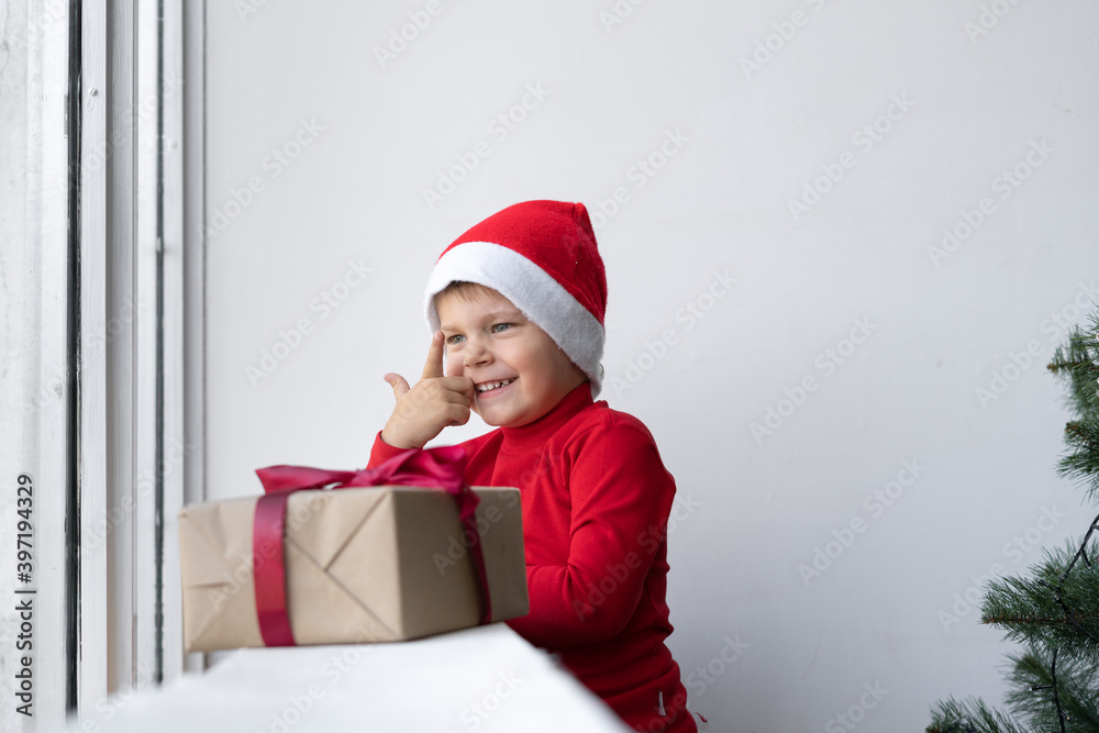 a cheerful little boy stands near the window wearing a santa claus hat and raises his index finger. a cheerful little boy stands near the window wearing a santa claus hat and raises his index finger.