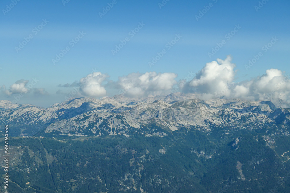 A panoramic view on a distant chain of Alps from another high peak. The massive chain is partially shrouded with clouds. Below there is lush green valley and a few cities in it. Austrian landscape A panoramic view on a distant chain of Alps from another high peak. The massive chain is partially shrouded with clouds. Below there is lush green valley and a few cities in it. Austrian landscape